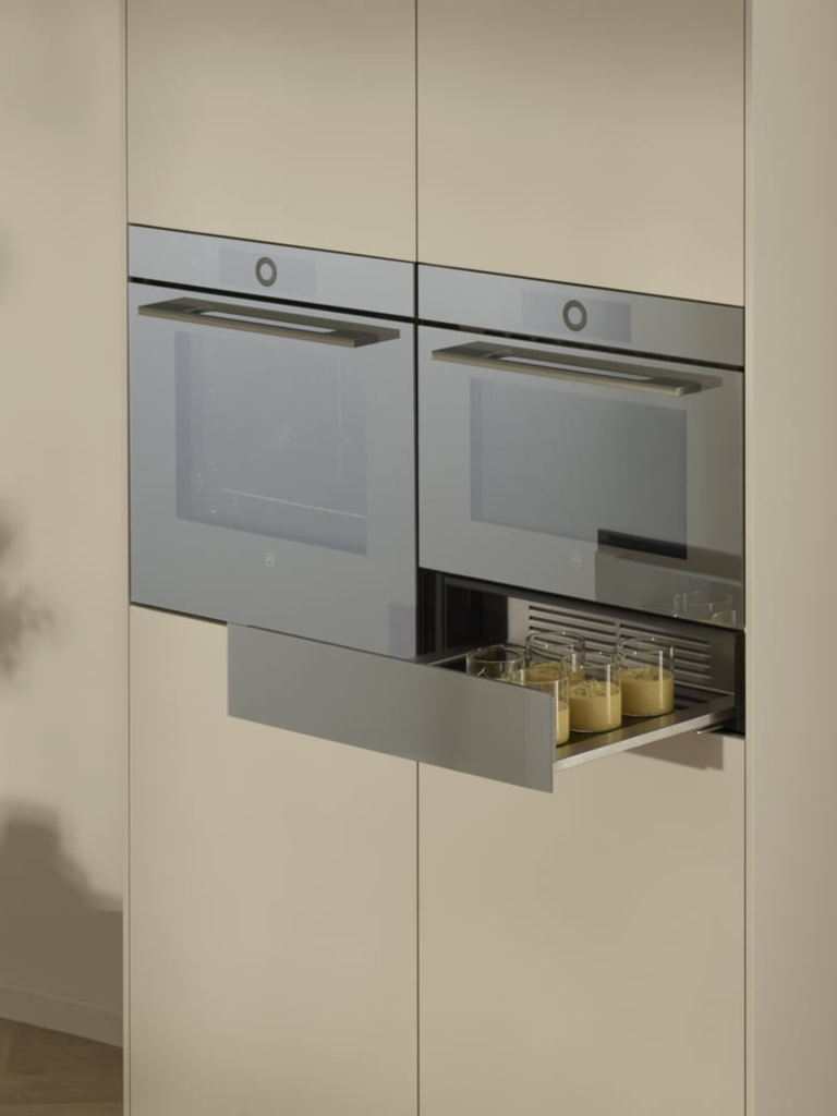 Built-in V-ZUG kitchen ovens with a warming drawer partially open, holding glass dishes inside a sleek modern cabinetry design
