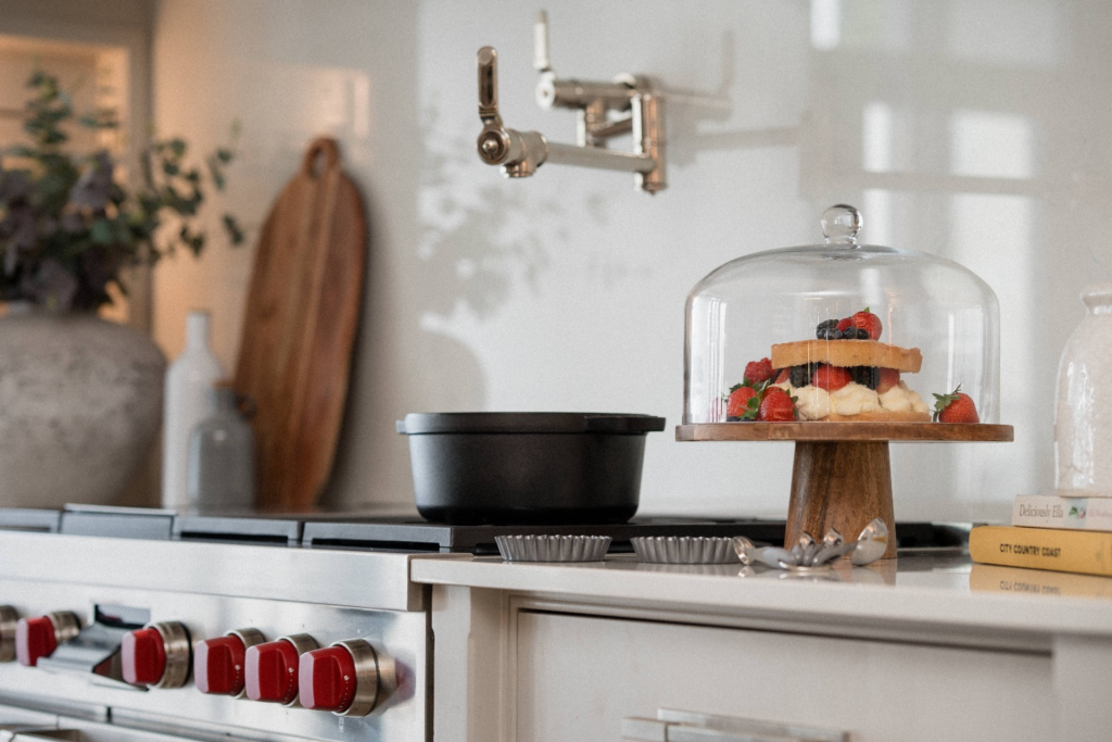 Close-up of a luxury kitchen with a Wolf cooker featuring red knobs, a pot filler tap, and a cake displayed under a glass dome