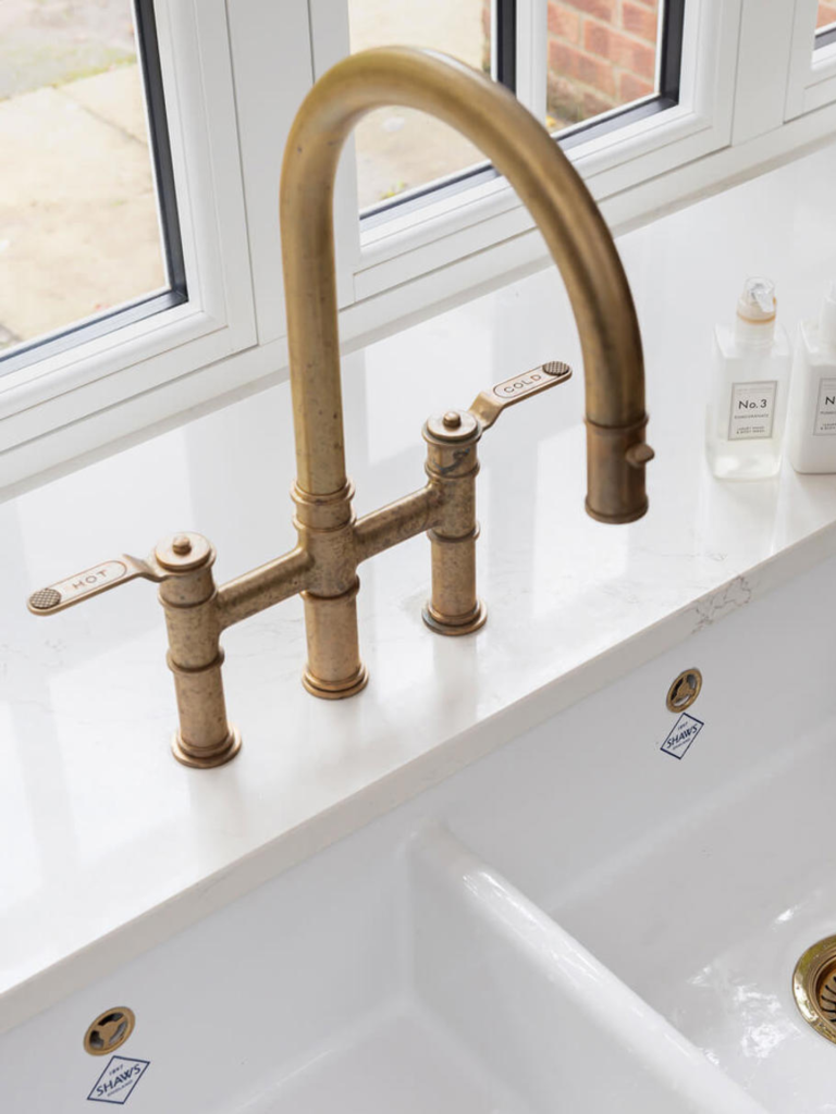 Close-up of a traditional brass kitchen tap with separate hot and cold levers, mounted on a white marble worktop above a white double Shaws ceramic sink with brass waste fittings