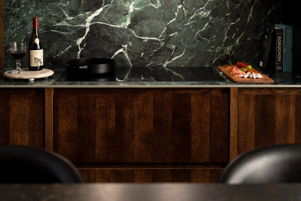 Elegant dark kitchen with green marble splashback, wooden cabinetry, a bottle of red wine, and a charcuterie board on the worktop