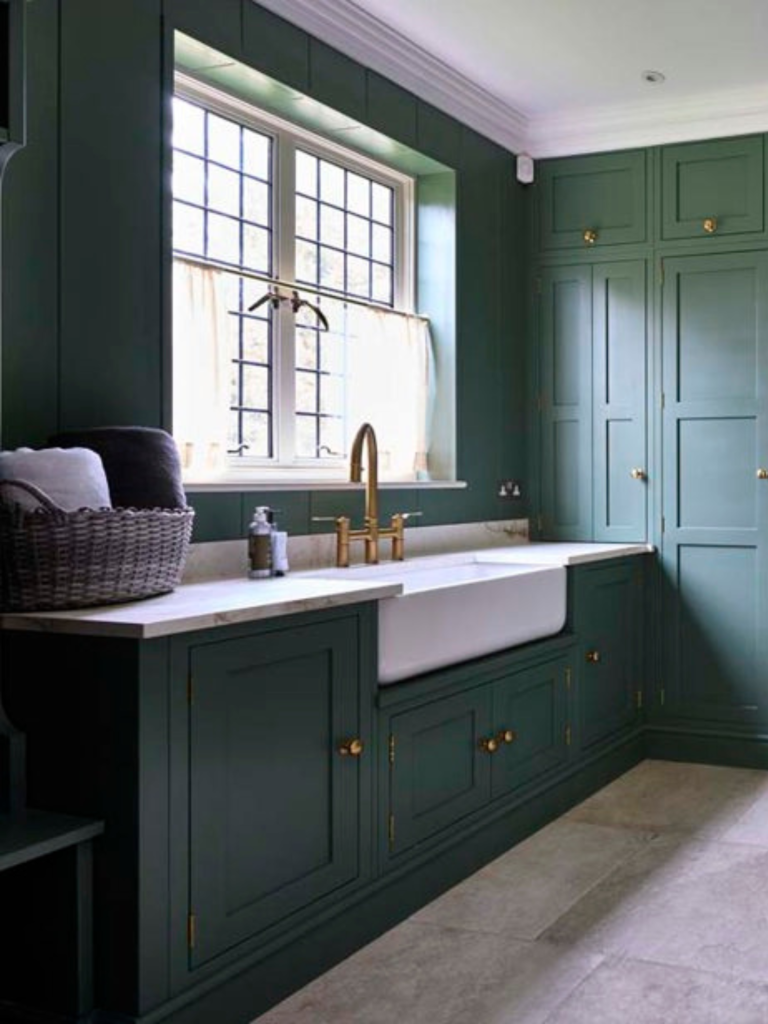 Traditional green shaker-style utility room with brass hardware, a large white Belfast sink, marble countertop, and a central brass tap beneath a window with divided panes and soft curtains