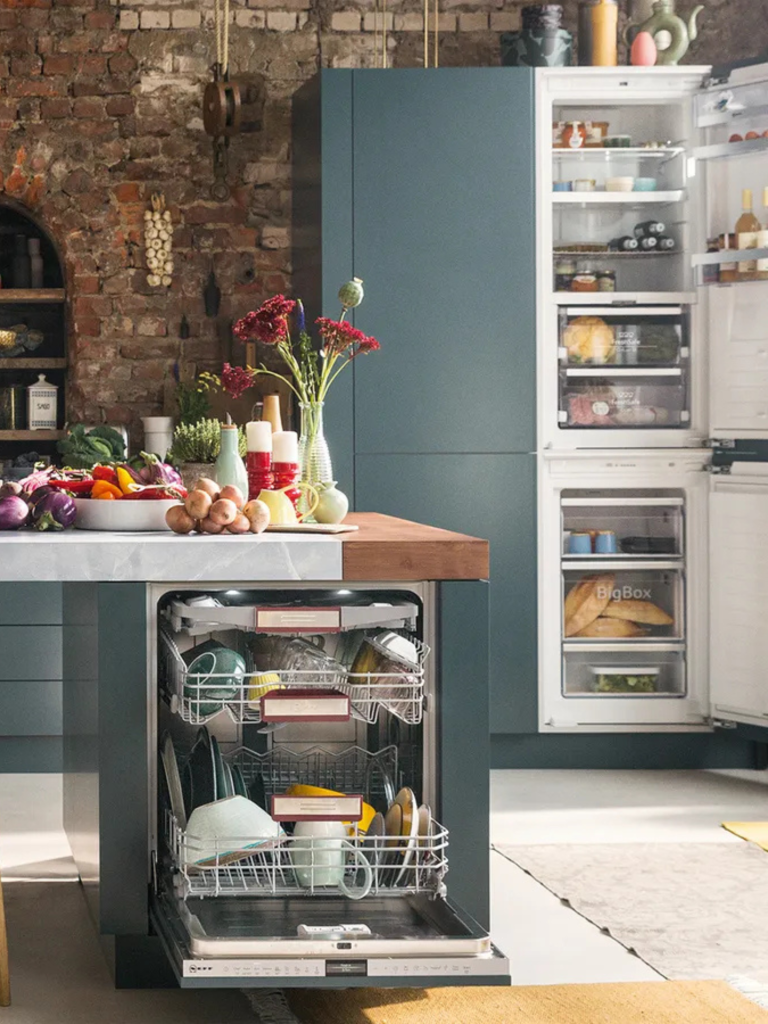 Open-plan kitchen with a fully loaded built-in dishwasher and an open fridge-freezer, set against a rustic brick wall and teal cabinetry