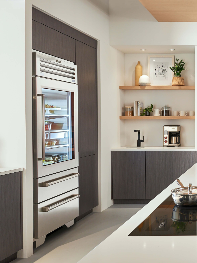 A Sub-Zero refrigerator with a glass door and stainless steel drawers integrated into a modern kitchen with dark wood cabinetry and minimalist open shelving