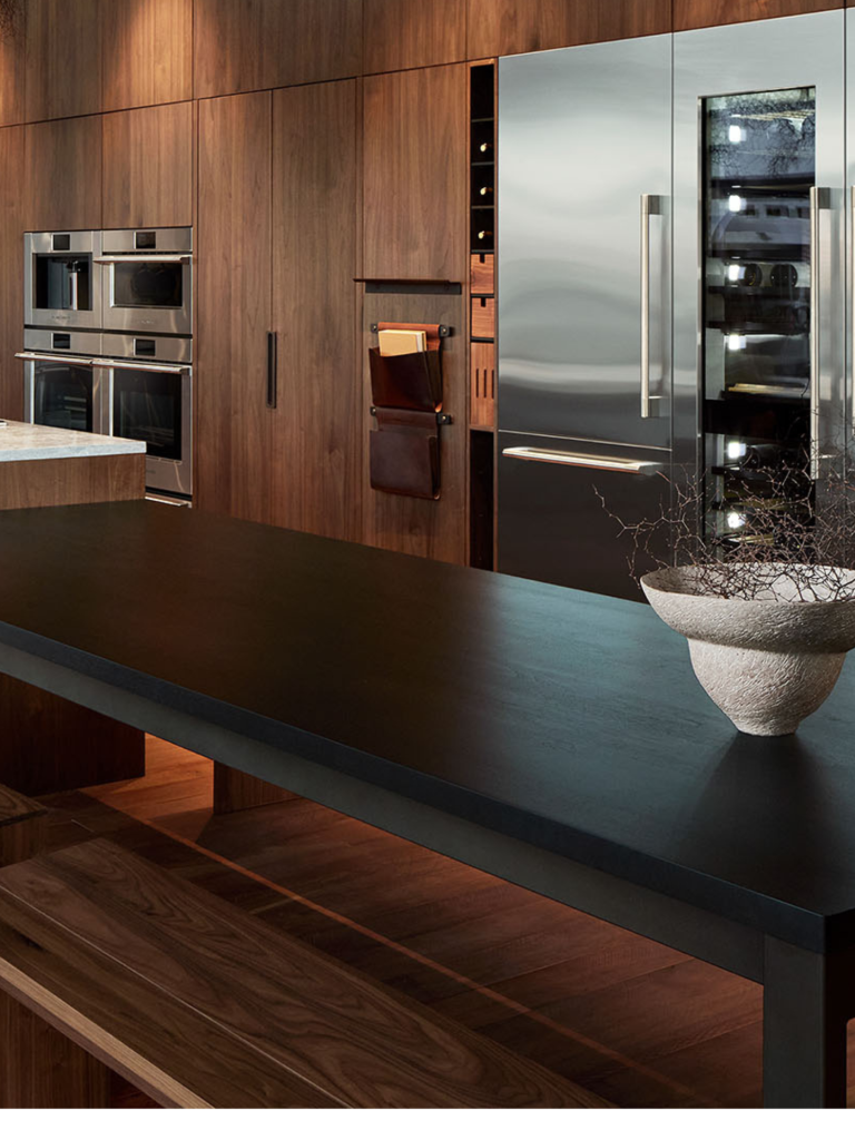 A modern kitchen with floor-to-ceiling walnut cabinetry featuring built-in stainless steel appliances, including ovens and a large Sub-Zero refrigerator with a glass-front wine column, set beside a sleek black island counter