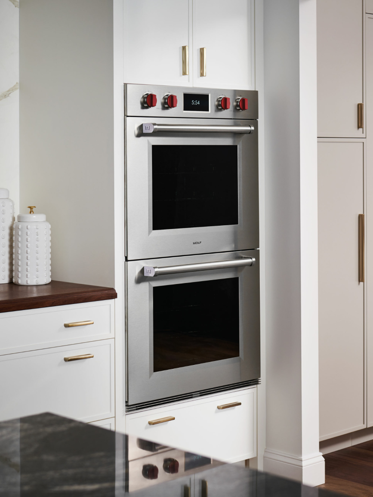 A pair of stainless-steel Wolf wall ovens with signature red knobs built into a white kitchen cabinet run, accented with brass hardware and a dark wood countertop