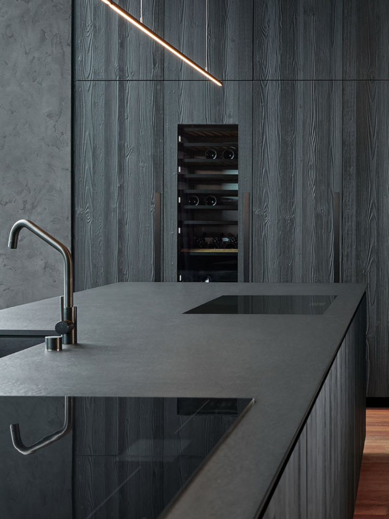 A dark, contemporary kitchen with textured charcoal cabinetry and a tall integrated wine fridge, paired with a large black island countertop and minimalist stainless steel tap