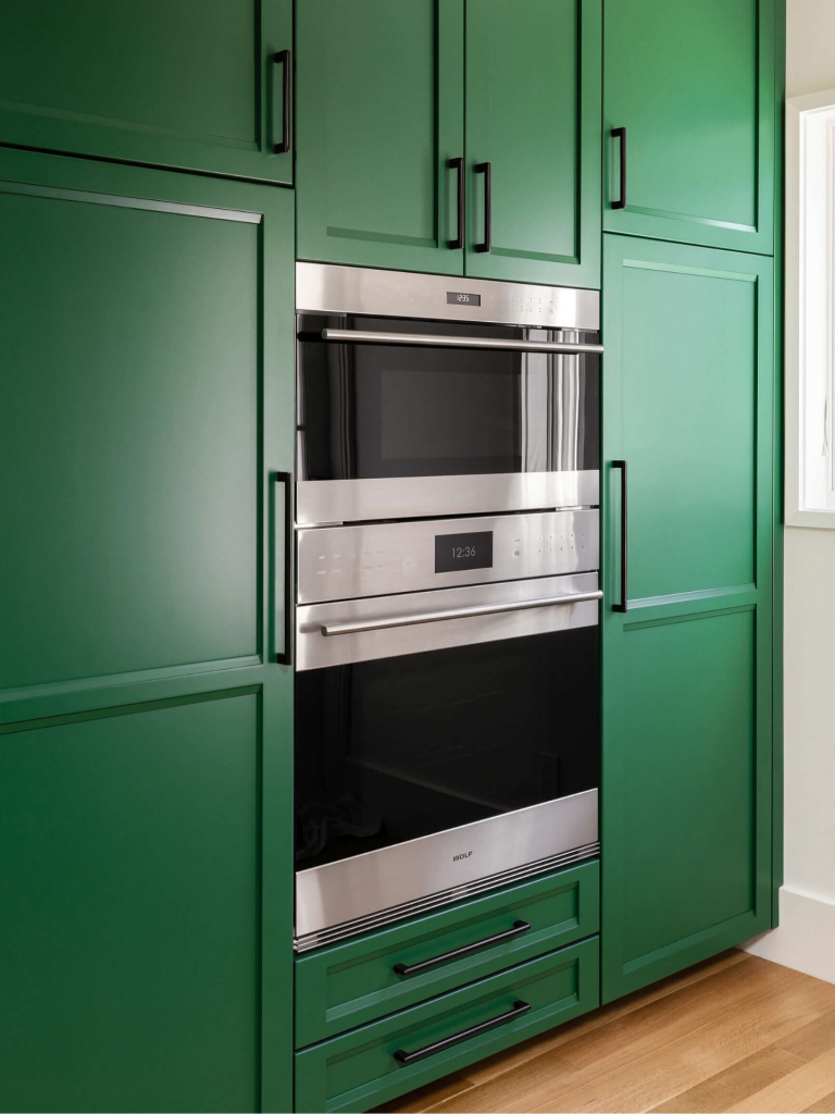A built-in Wolf microwave and oven combination seamlessly integrated into bold green shaker-style kitchen cabinetry with black handles and natural wood flooring