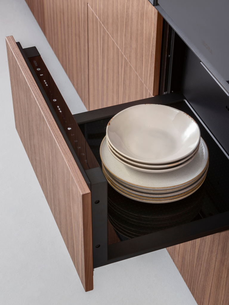 Open warming drawer with stacked ceramic plates inside a modern wood-panelled kitchen
