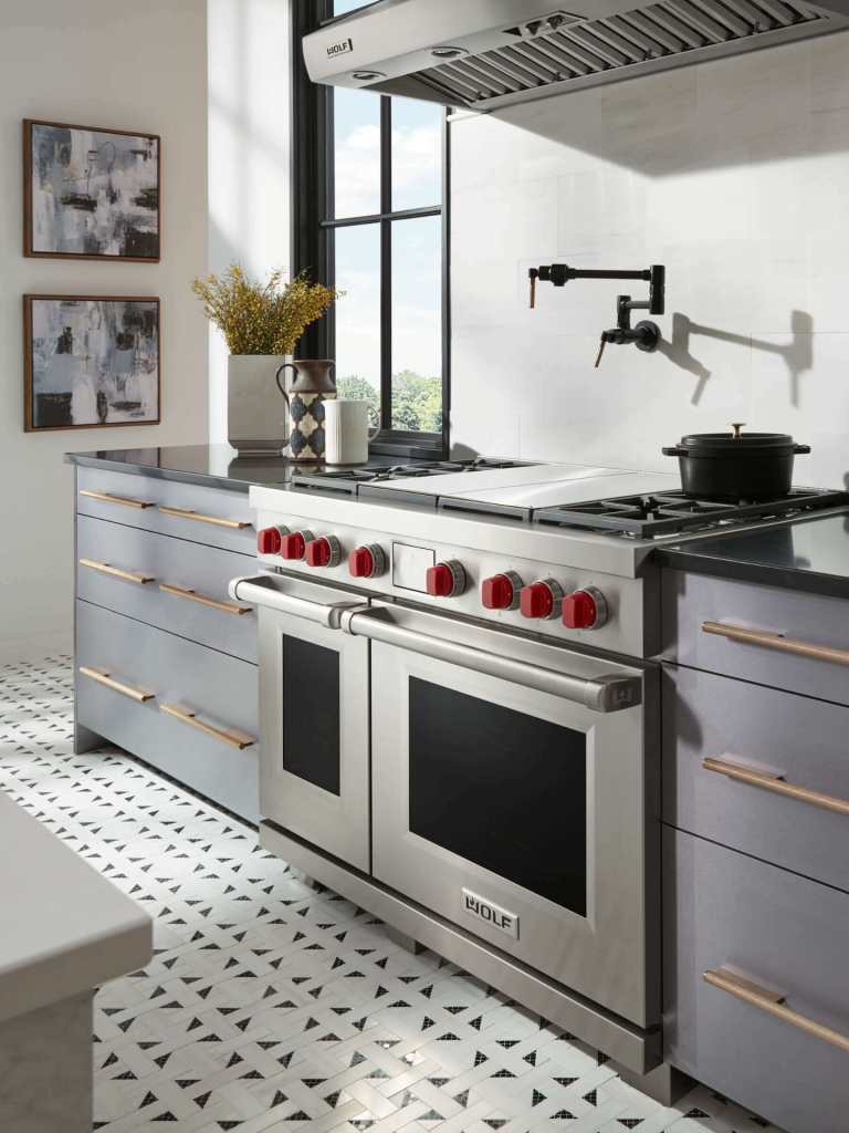 A stainless-steel Wolf dual-fuel range with signature red knobs installed in a bright, modern kitchen featuring grey cabinetry, black countertops, and patterned white floor tiles beneath a large industrial-style cooker hood