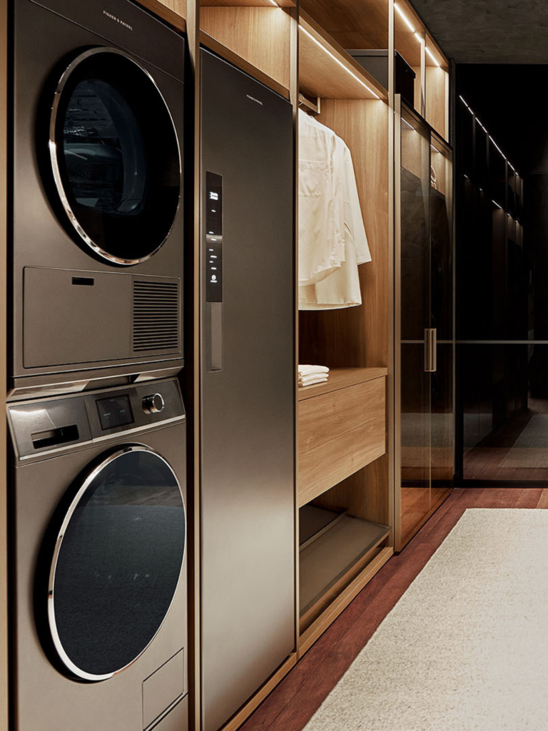 A luxury laundry space with Fisher & Paykel appliances, including a stacked washer and dryer next to a tall drying cabinet, all integrated within warm wood cabinetry and softly lit shelving