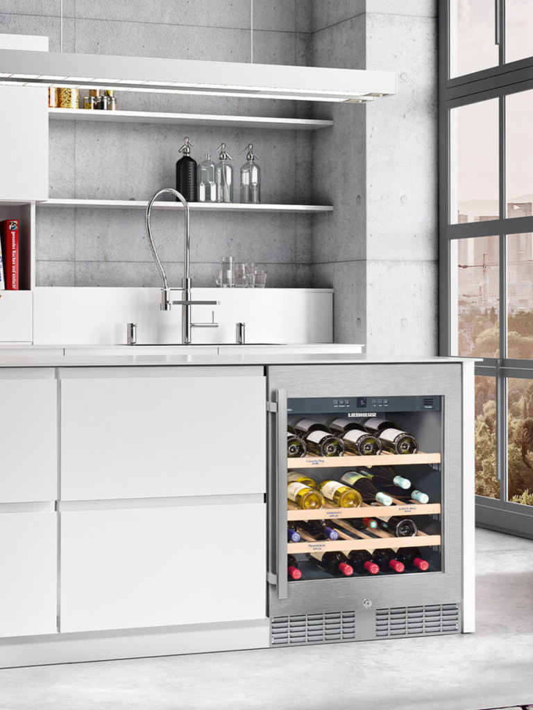 Built-in Liebherr undercounter wine cooler integrated into a sleek modern white kitchen with open shelving and stainless-steel accents