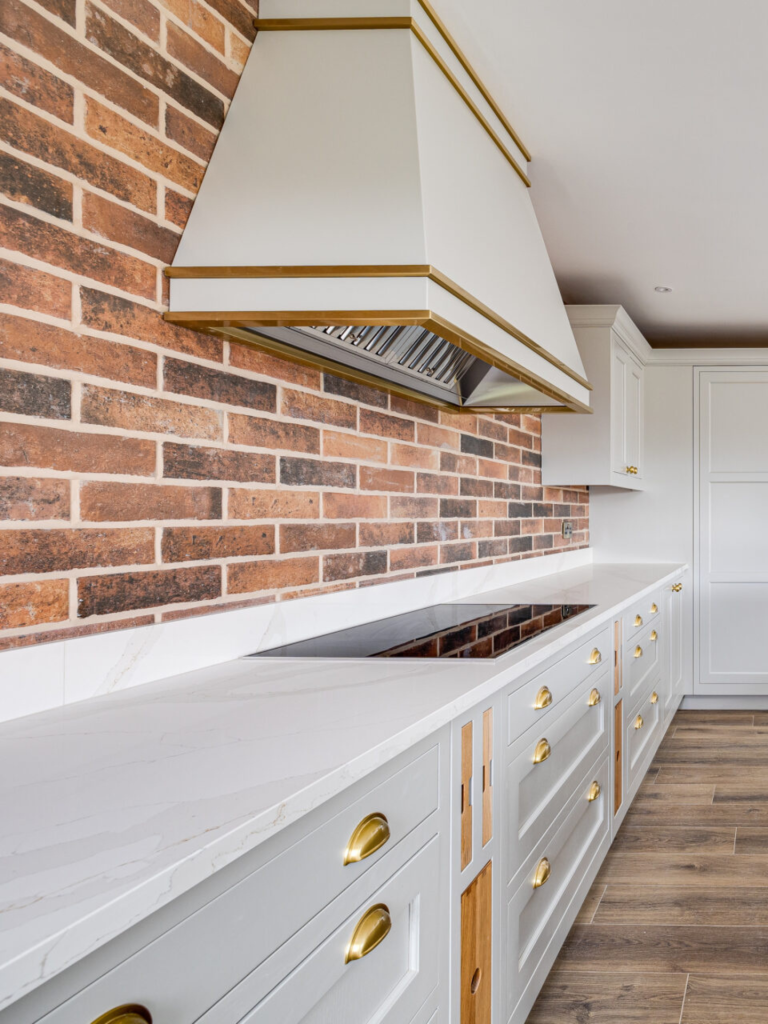 Bespoke Westin white and gold kitchen canopy hood set against a red brick backsplash, featuring marble countertops and shaker cabinetry with gold cup handles