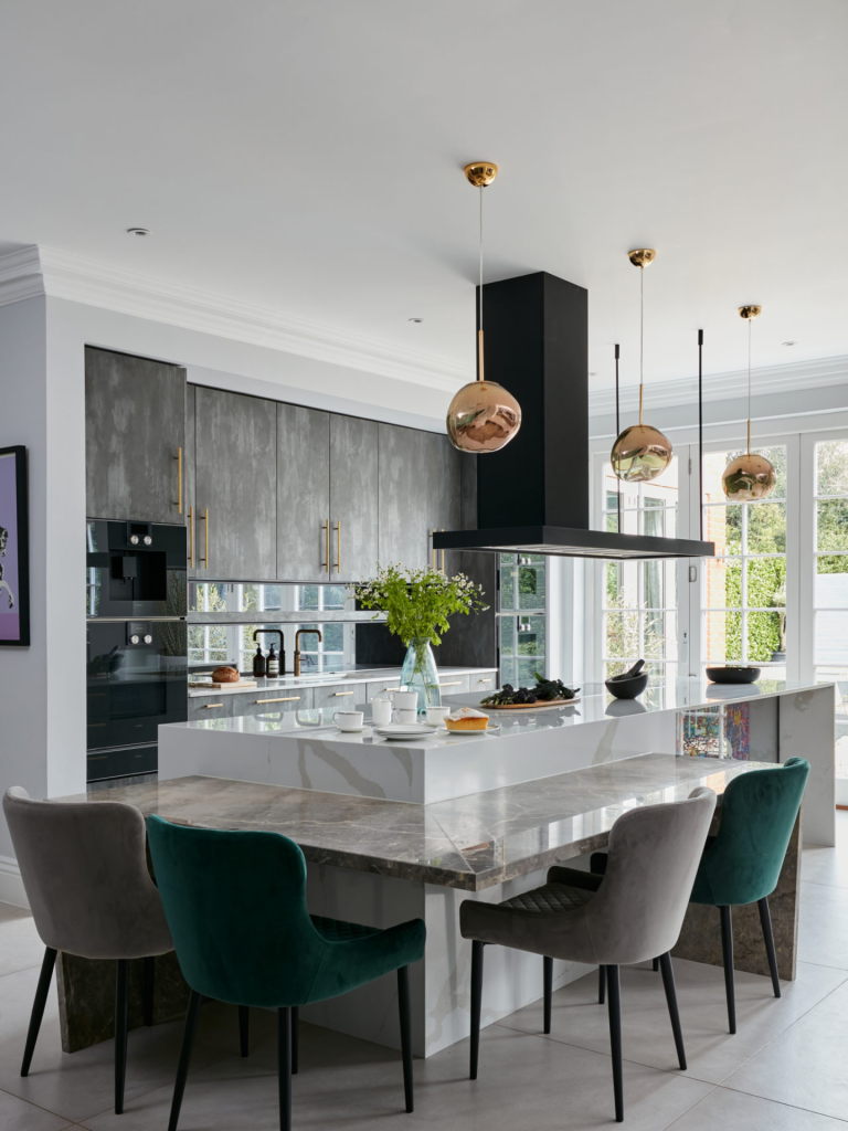 Modern open-plan kitchen with grey textured cabinets, marble waterfall island, and gold pendant lights above a built-in dining extension with velvet chairs