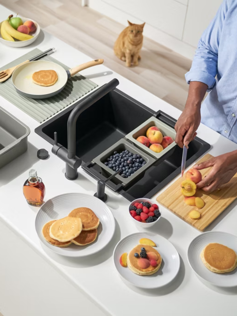 A modern kitchen scene with a black multifunctional sink workstation filled with blueberries and peaches, surrounded by plates of pancakes and fresh fruit, with a person slicing peaches and a cat sitting in the background