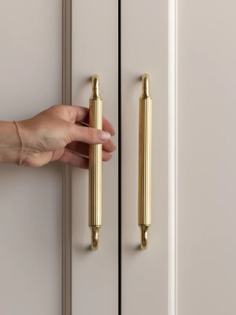 Close-up of a hand opening tall cabinet doors fitted with long, fluted brass handles in a soft, neutral-toned kitchen