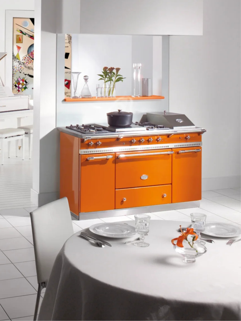A bright orange Lacanche range cooker positioned in a minimalist white kitchen, with matching silver details and a contemporary dining table in the foreground