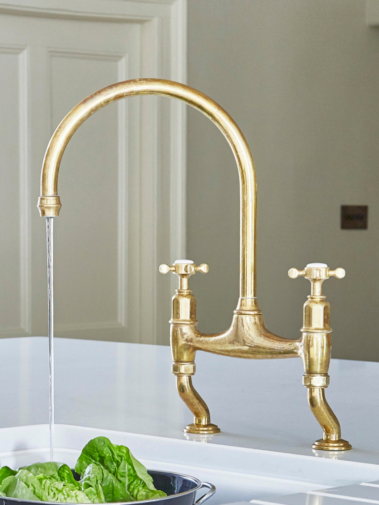 Close-up of a traditional brass kitchen faucet with a high curved spout and dual cross handles, mounted on a white countertop with water running into the sink