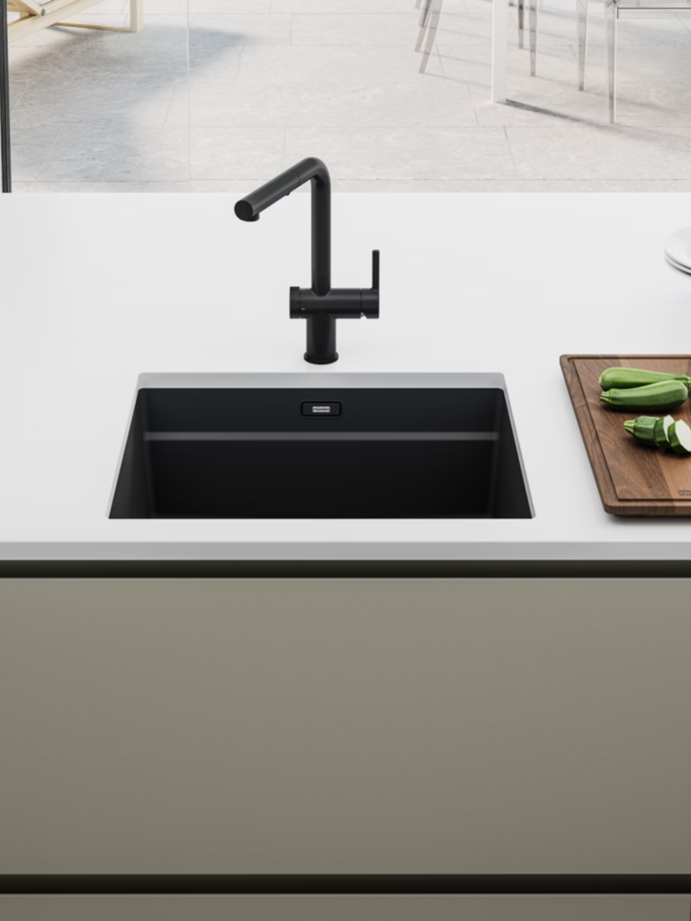 A minimalist kitchen sink setup featuring a matte black Franke tap and matching black sink bowl set into a white countertop, with chopped vegetables on a wooden board nearby