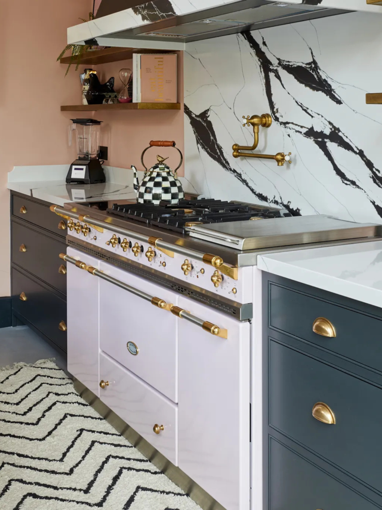 A white and gold Lacanche range cooker integrated into a modern kitchen, featuring brass accents, marble-effect backsplash, and surrounding dark cabinetry
