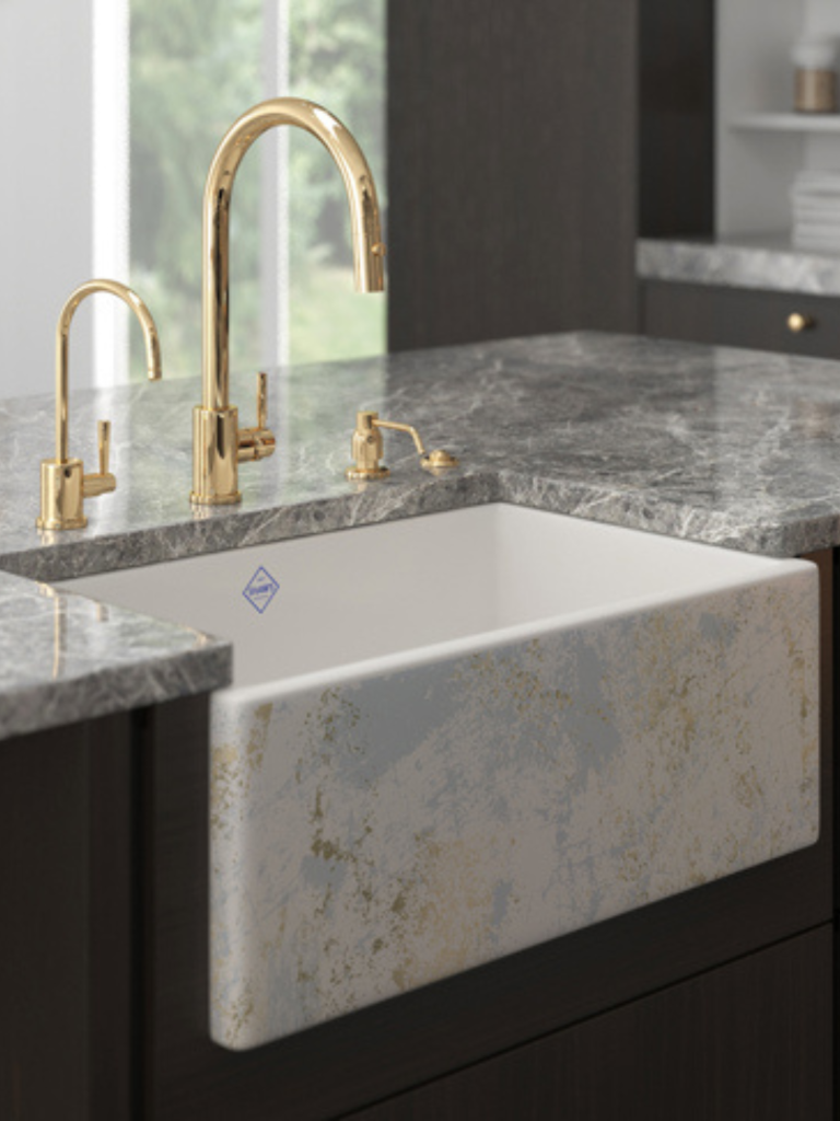 Gold kitchen tap with a high curved spout mounted on a grey marble countertop, paired with a decorative front-apron ceramic sink featuring a subtle gold and grey marbled pattern
