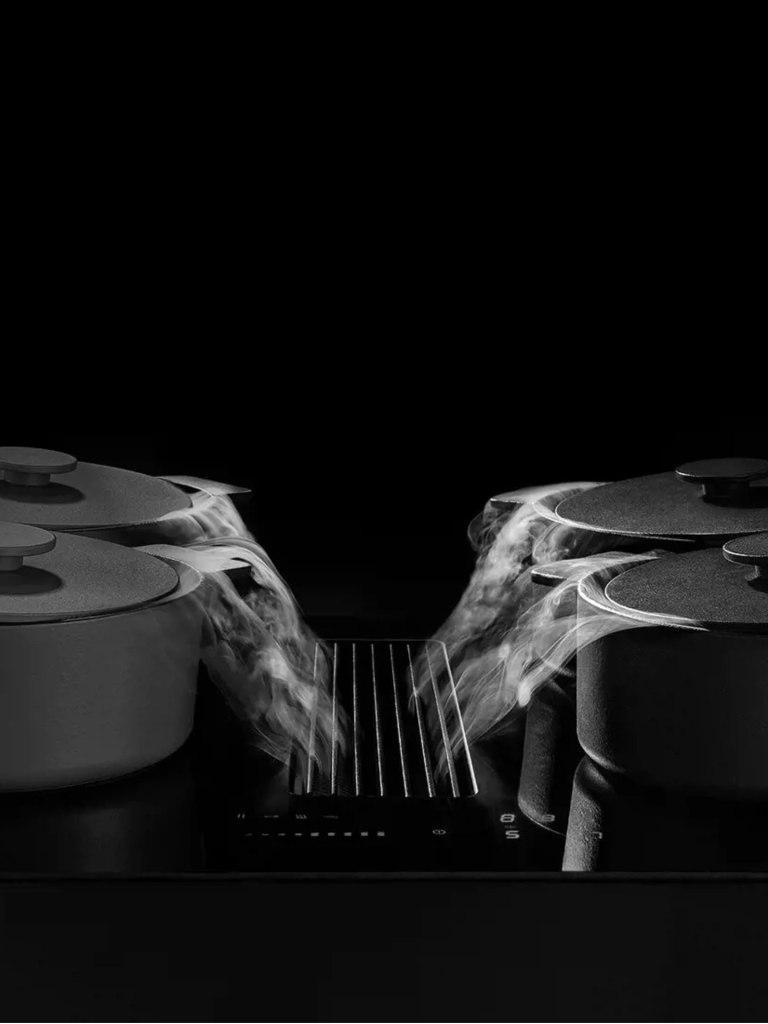 A sleek Novy black cooktop with integrated downdraft extraction pulling steam from two cooking pots, creating a dramatic high-contrast visual against a dark background