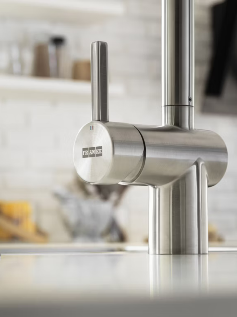 A close-up of a stainless steel Franke kitchen tap, showcasing its sleek cylindrical design and brushed metal finish against a soft-focus kitchen backdrop