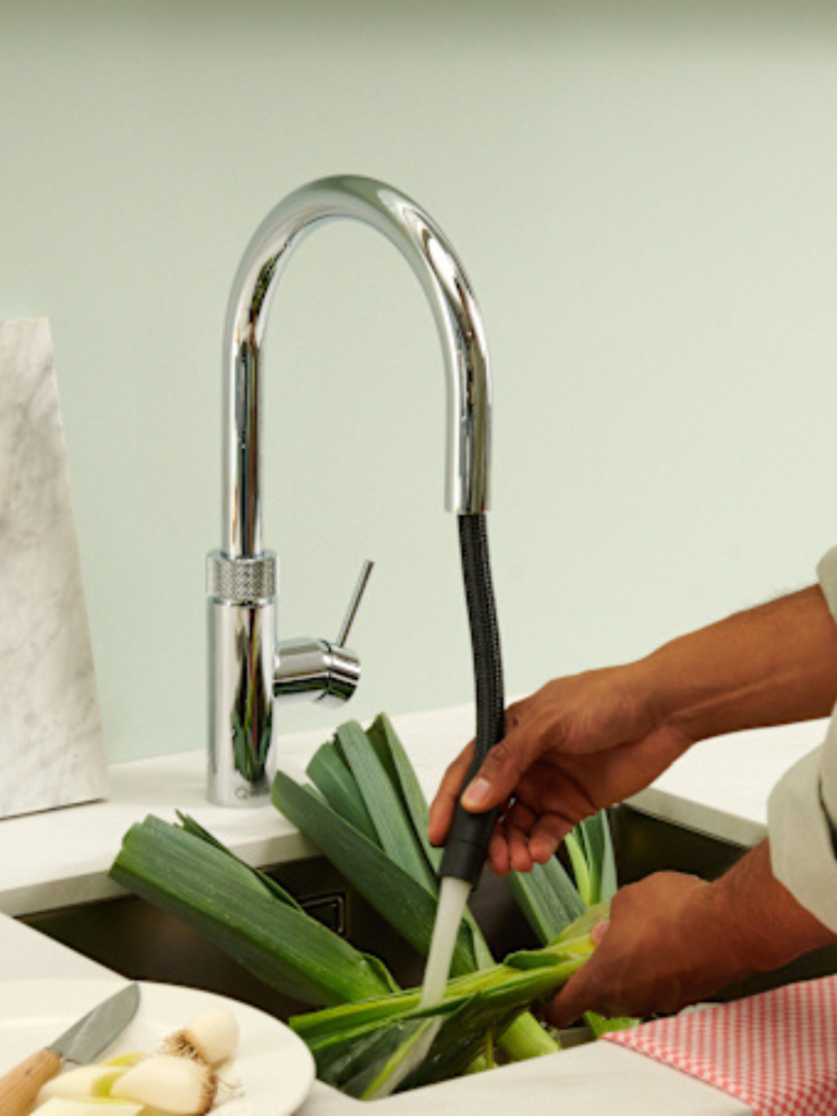 Chrome pull-out kitchen tap being used to rinse fresh leeks in a stainless steel sink, with a light green wall and white countertop