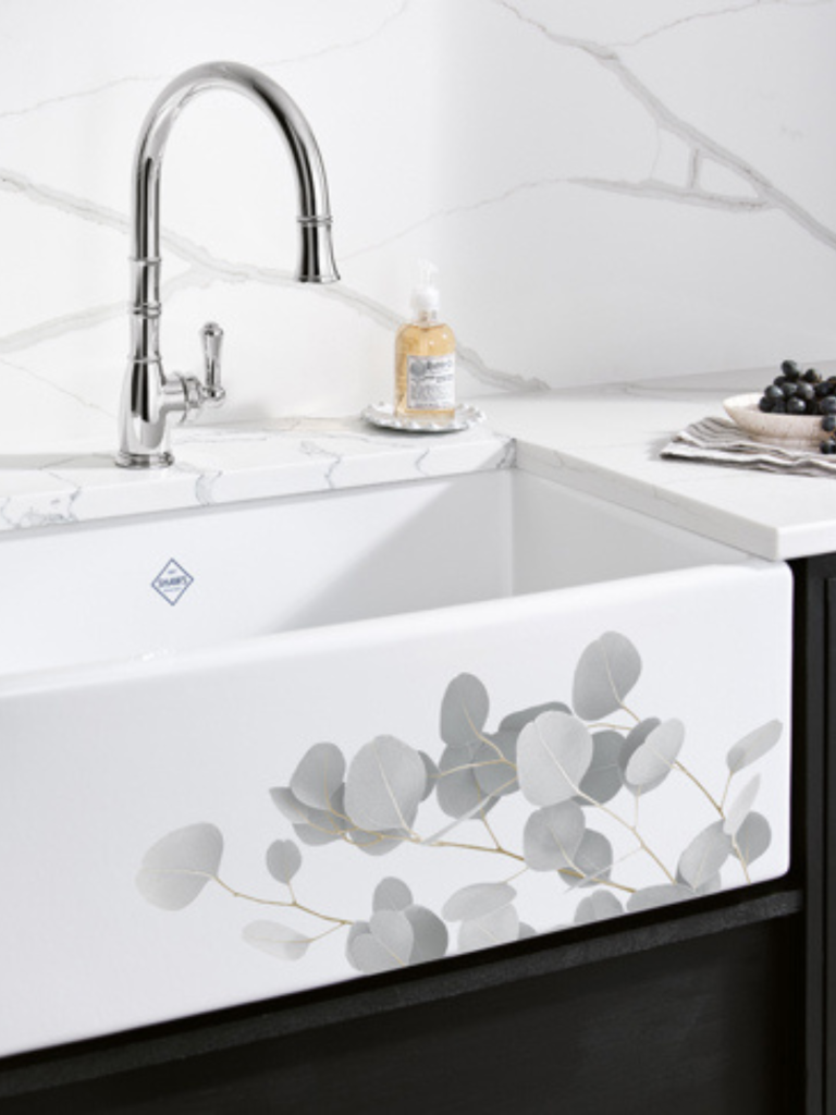 Chrome kitchen tap with a curved spout and single lever, installed above a white ceramic apron-front sink decorated with a grey eucalyptus leaf design