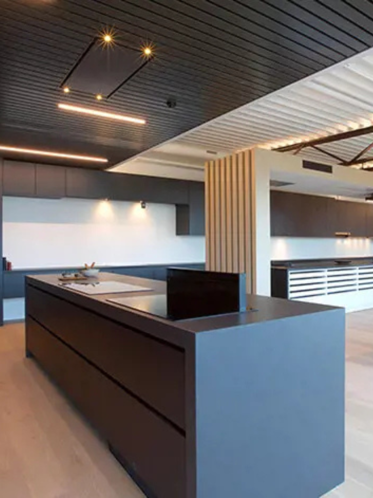 A modern kitchen featuring a minimalist island with a concealed downdraft extractor partially raised, complemented by a dark slatted ceiling with integrated lighting