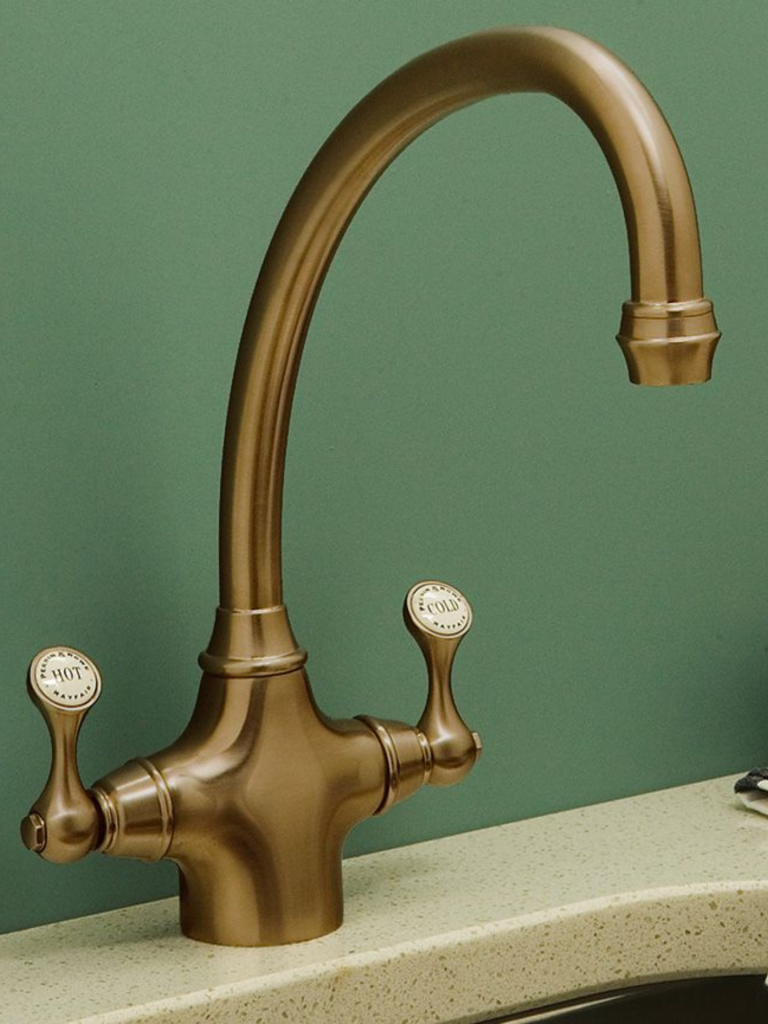 Vintage-style bronze kitchen faucet with a high arching spout and labeled hot and cold lever handles, installed on a light-colored countertop against a green wall