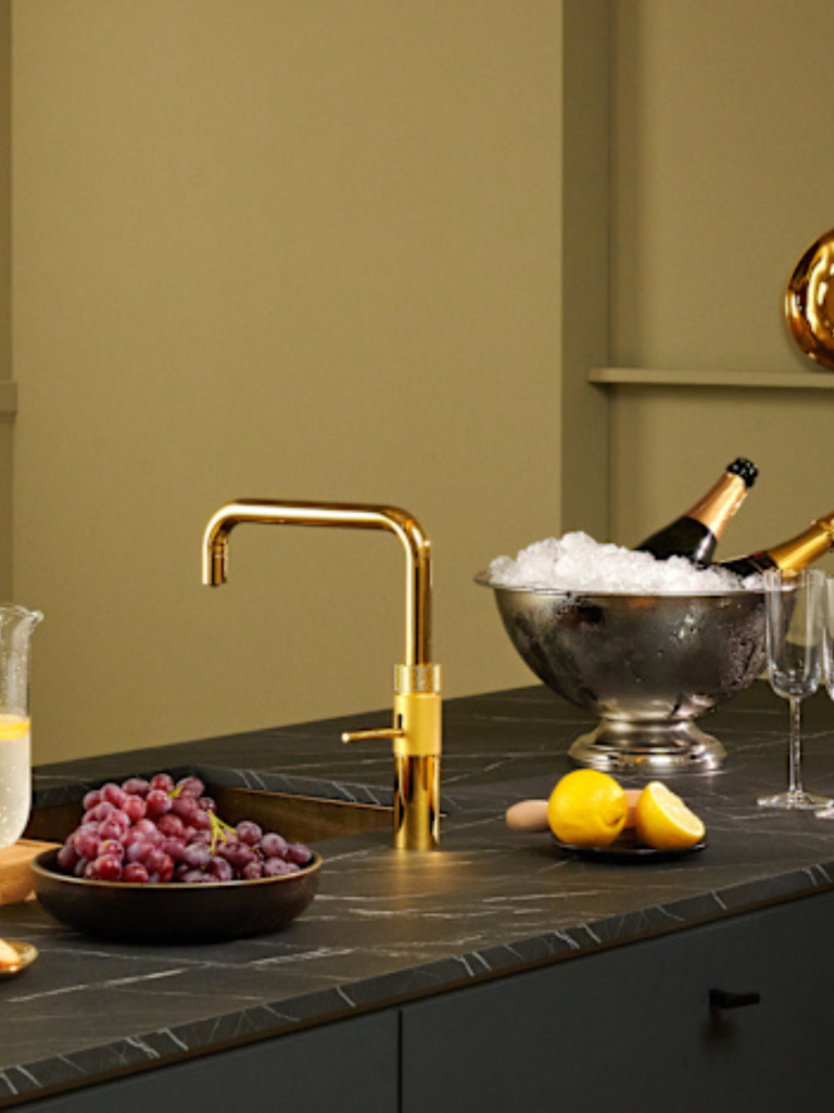 Gold kitchen tap installed on a dark marble countertop, surrounded by grapes, lemons, and a champagne bowl filled with ice