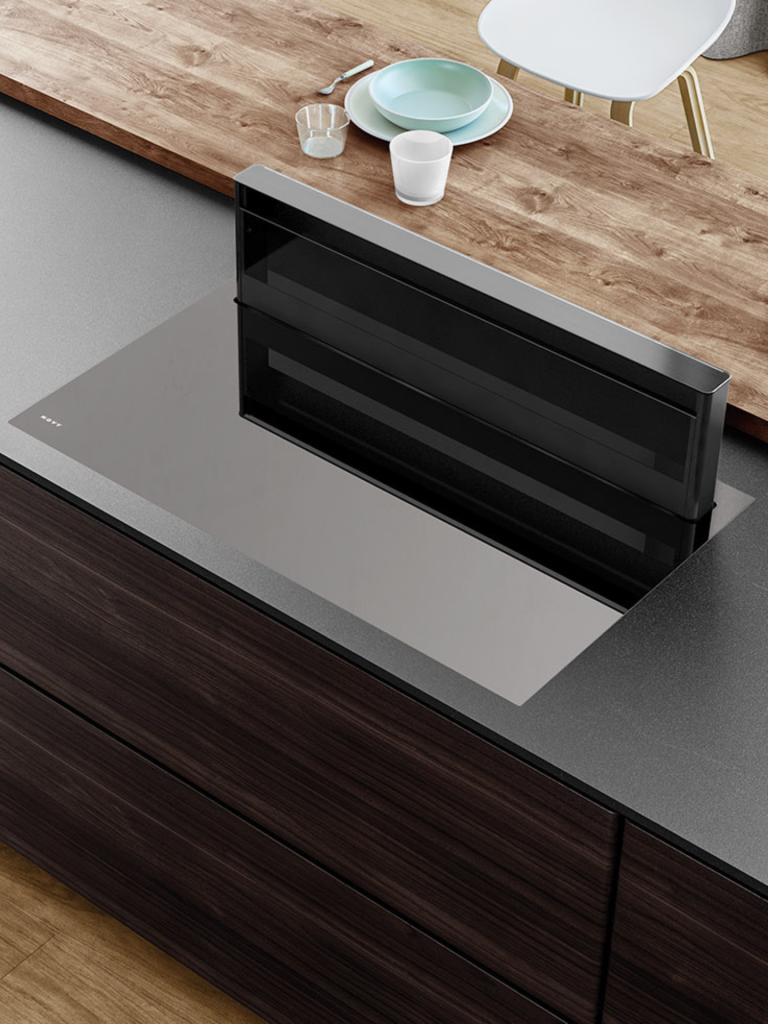 A close-up view of a contemporary cooktop with a fully raised downdraft extractor set into a dark wood kitchen island with a contrasting wood breakfast bar