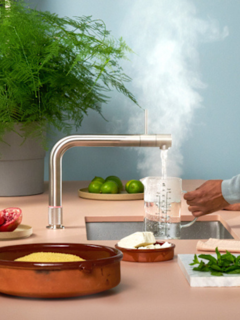 Modern kitchen tap dispensing steaming hot water into a glass jug on a pastel countertop, with fresh ingredients and leafy green plants in the background