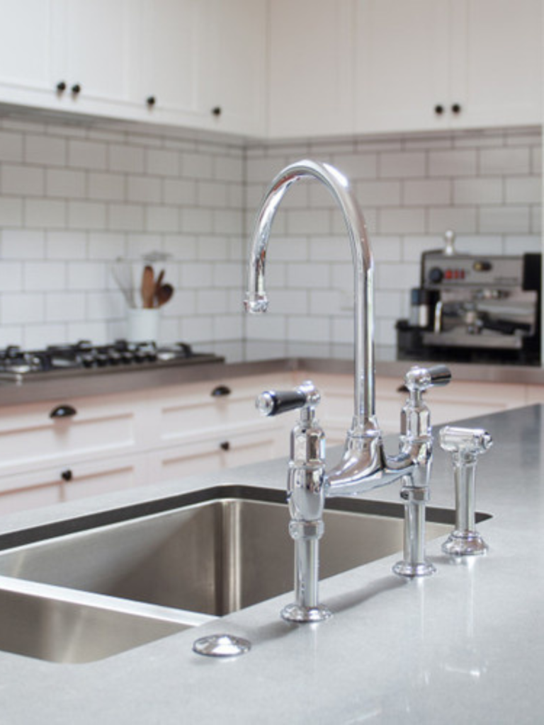 Polished chrome kitchen faucet with a tall curved spout and dual lever handles, alongside a side sprayer, mounted on a gray countertop in a white subway-tile kitchen