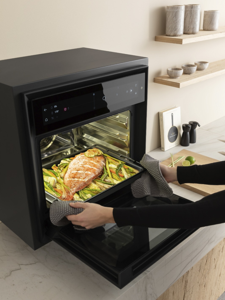 Person removing a tray of baked salmon and vegetables from a modern V-Zug oven in a light, minimalist kitchen setting