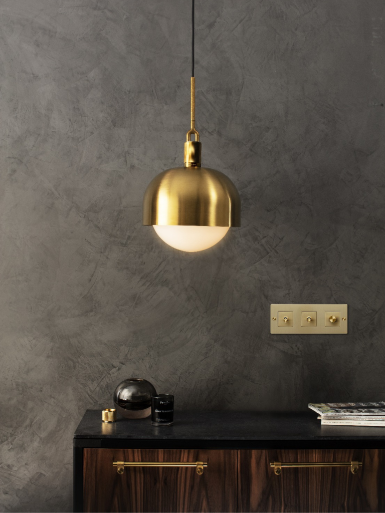 Elegant brass pendant light above a walnut cabinet with matching hardware and brass light switches on a textured grey wall