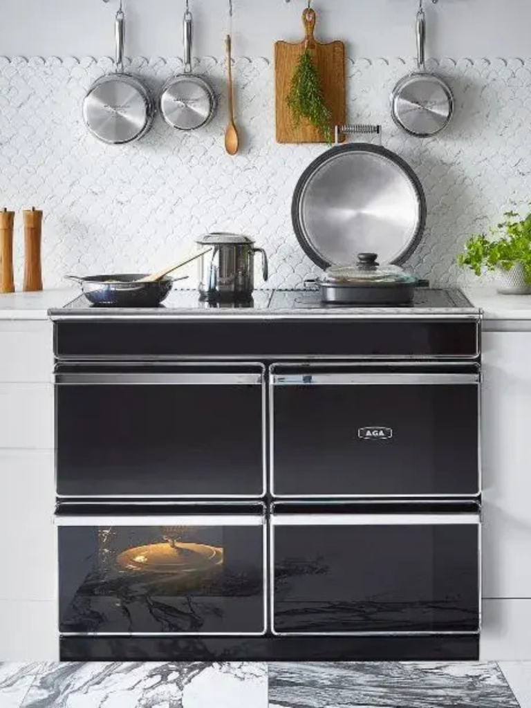 A black AGA cooker in a modern kitchen, with multiple ovens, a hotplate open, and stainless-steel pans hanging above a tiled backsplash