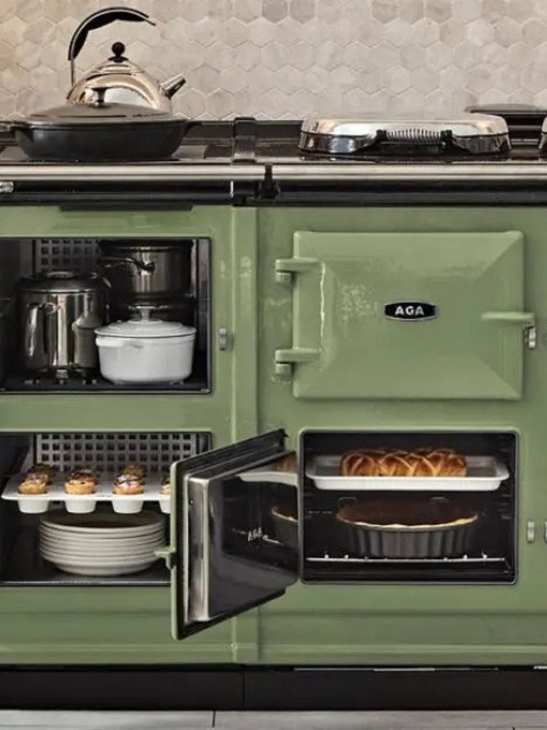 A green AGA range cooker with multiple ovens open, showing baked goods and cookware inside, with pans and a kettle on the hotplates above