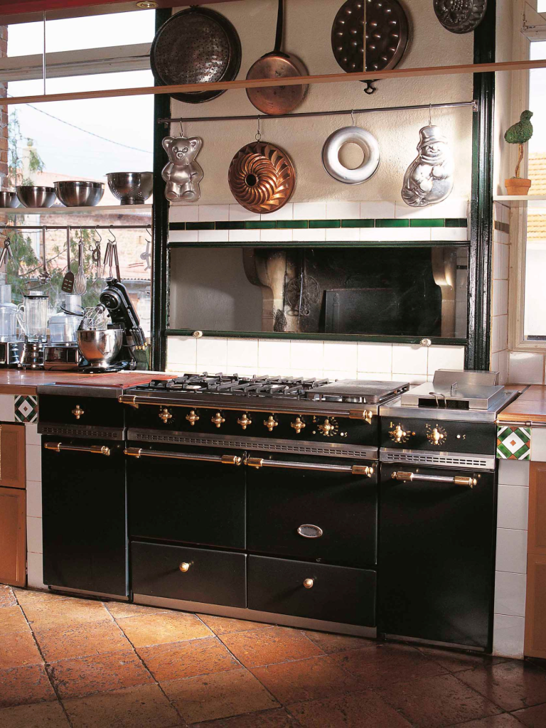 A large black La Cornue-style range cooker with brass knobs and handles installed in a rustic kitchen, surrounded by hanging copper and metal cookware and tiled countertops